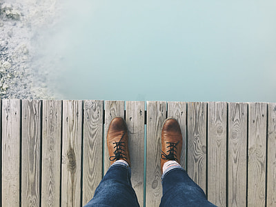 person wearing blue denim pants and brown casual shoes standing on gray dock above water thumbnail