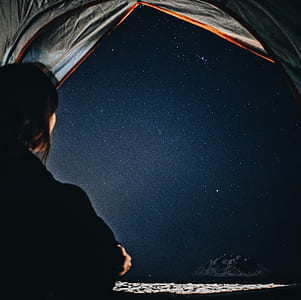 person wearing black coat sitting inside gray camping tent with view of seashore during nighttime thumbnail