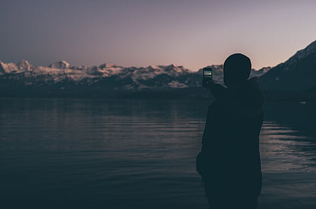 person standing beside body of water near on mountain alps while holding smartphone thumbnail