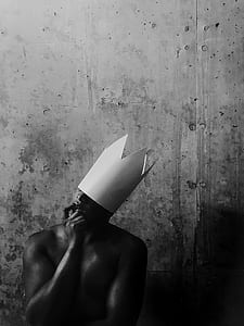 man biting his right index finger while wearing paper made crown standing near wall thumbnail