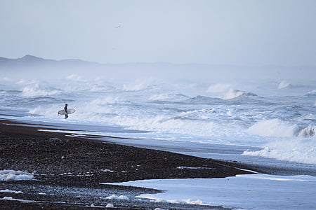 person carrying surfboard standing on body of water thumbnail
