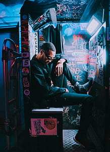 man in black jacket and blue jeans sitting on white bench with one foot raised inside a room with graffiti thumbnail