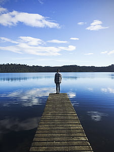 man standing on wooden dock facing lake during daytime thumbnail