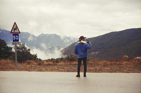 person wearing blue jacket carrying cruiser board while standing on road thumbnail