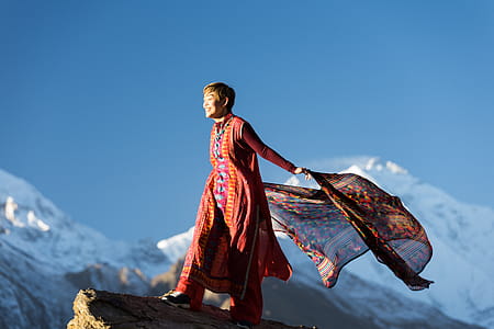 Woman in Orange Dress Standing on Top of Rock Cliff Holding Scarf Near Mountain Covered With Snow thumbnail