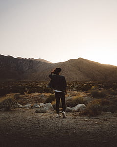 person wearing blue denim jacket and black hat standing in front of rock formations during golden hour thumbnail