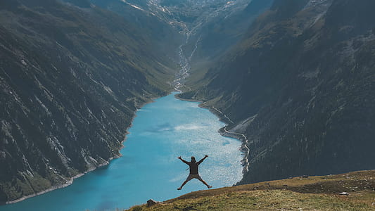 man jumping near calm body of water between two green mountain during daytime thumbnail