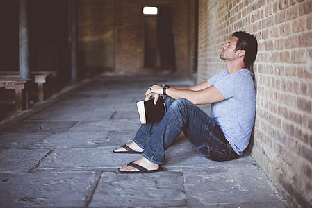 man wearing gray shirt holding book sitting beside brick wall thumbnail