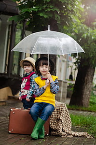 girl standing near boy sitting on brown case holding clear umbrella thumbnail