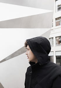 man in black hoodie standing near building wall during daytime thumbnail