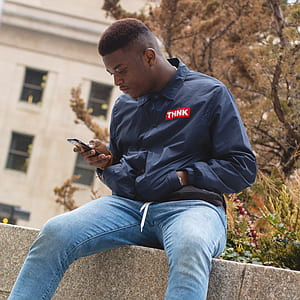 man wearing blue jacket sitting on concrete pavement while using phone thumbnail