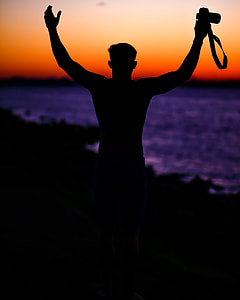 silhouette photography of man holding camera facing body of water thumbnail