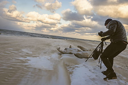Man in Black Bubble Jacket Walking on Seashore While Holding Black Camera Tripod thumbnail