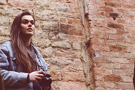woman wearing blue denim jacket standing beside bricked building while holding camera thumbnail