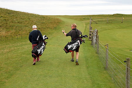 two men walking on grass field carrying golf bags thumbnail