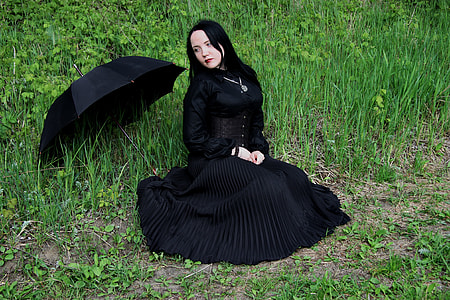 woman in black long sleeve long dress beside black umbrella and sitting on green grass  field thumbnail