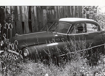 grayscale photo of classic car parked beside wooden wall thumbnail