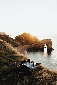 man laying on brown grass near cliff and body of water thumbnail