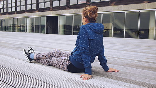 woman wearing blue hooded jacket, beige leggings and black-and-white lace-up low-top sneakers sits on beige wooden floor near clear glass door beige concrete building during daytime thumbnail