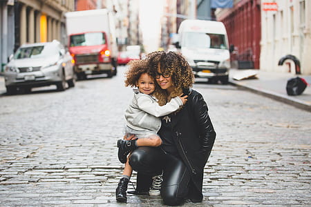mother and child kneeling on grey concrete road between buildings and vehicles during daytime thumbnail