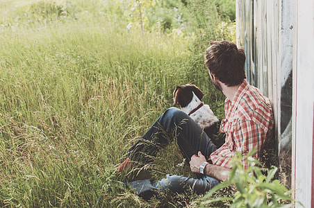 man in red,white,blue plaid sports shirt sitting on grass field beside black and white dog during daytime thumbnail
