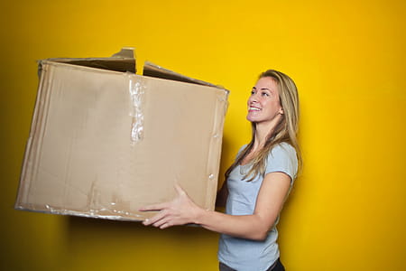Young Woman Holding Brown Cardboard Box Against Yellow Wall thumbnail