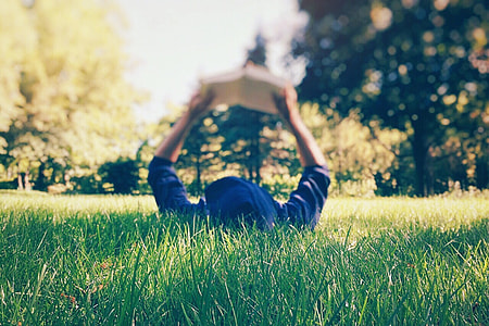 person in blue hoodie holding book while lying on green grass during daytime thumbnail