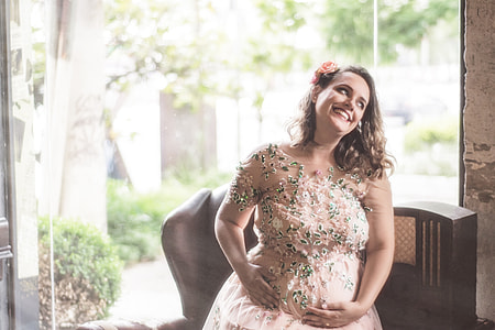 photo of pregnant woman in beige floral dress sitting on brown wooden sofa smiling thumbnail