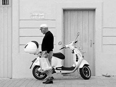 grayscale photography of man standing near motor scooter thumbnail