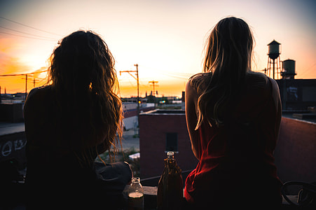 two women sitting near bottle in front of brown building during sunset thumbnail