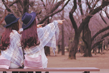 selective focus photography of two women wearing blue fedora hats pointing at the right side sitting in brown wooden bench chair during daytime thumbnail
