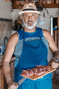 Man in Blue Kitchen Apron Holding Knife and Chopping Board With Sliced Meat thumbnail