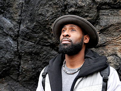 photography of man in white and black hoodie standing near black rock formation with his head held high during daytime thumbnail