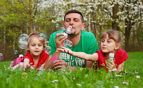 man with two girls playing with bubbles on green grass field during daytime thumbnail