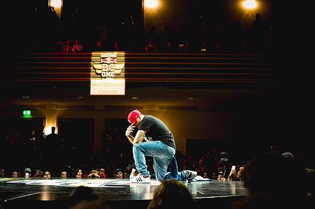 man wearing black t-shirt, red cap and blue denim jeans posing on stage in front of people thumbnail