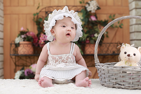 baby in white dress and head band beside wicker brown basket on white fur surface thumbnail