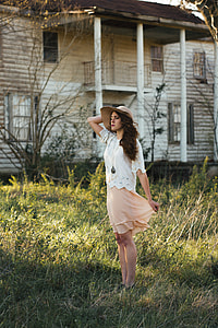 woman wearing gray sun hat, white shirt and peach-colored skirt standing in green grass and white wooden house thumbnail