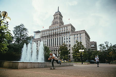 Young Caucasian skateboarders ride past a fountain in front of a building thumbnail