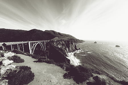 grayscale photo of concrete bridge connecting two mountains near body of water thumbnail