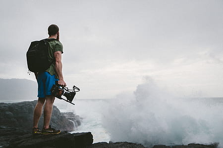 man carrying a backpack and video recorder on rock formation near body of water thumbnail