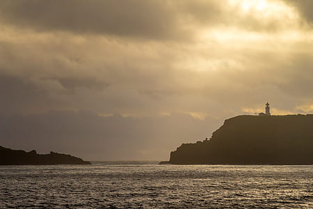 silhouette photo of lighthouse on mountain across body of water during golden hour thumbnail