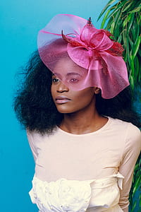 woman wearing pink fascinator and peach-colored scoop-neck long-sleeved shirt behind tall green plant in teal background thumbnail