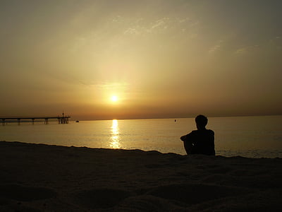 silhouette of man sitting on sea shore watching sunset thumbnail