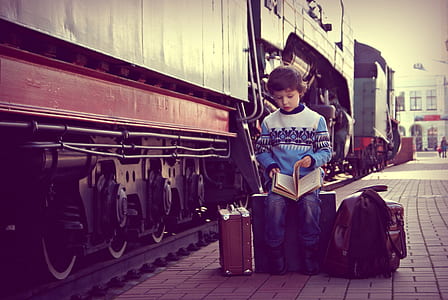 boy opening book page while sitting on luggage beside train outdoors thumbnail