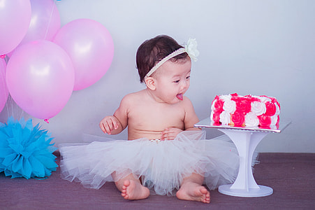 baby in white tutu skirt near white and red fondant cake and pink balloons thumbnail