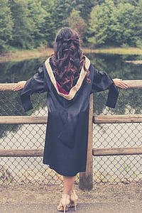 woman in black graduation gown standing near brown wooden fence thumbnail