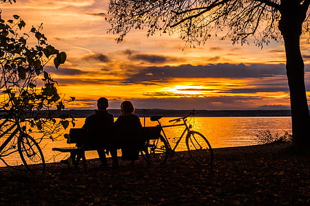 silhouette of couple sitting on bench near body of water during golden hour thumbnail