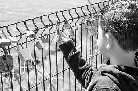 grayscale photo of boy holding padlock in metal fence thumbnail
