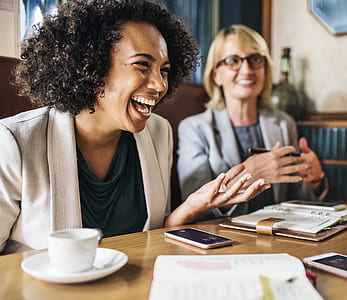 laughing woman wearing white blazer sitting beside woman wearing grey notched-lapel blazer and black framed eyeglasses thumbnail