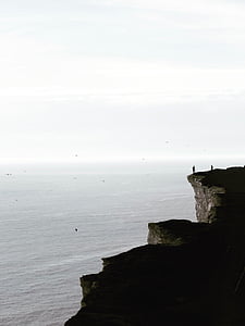 two people near the edge of the cliff near birds flying above the sea thumbnail